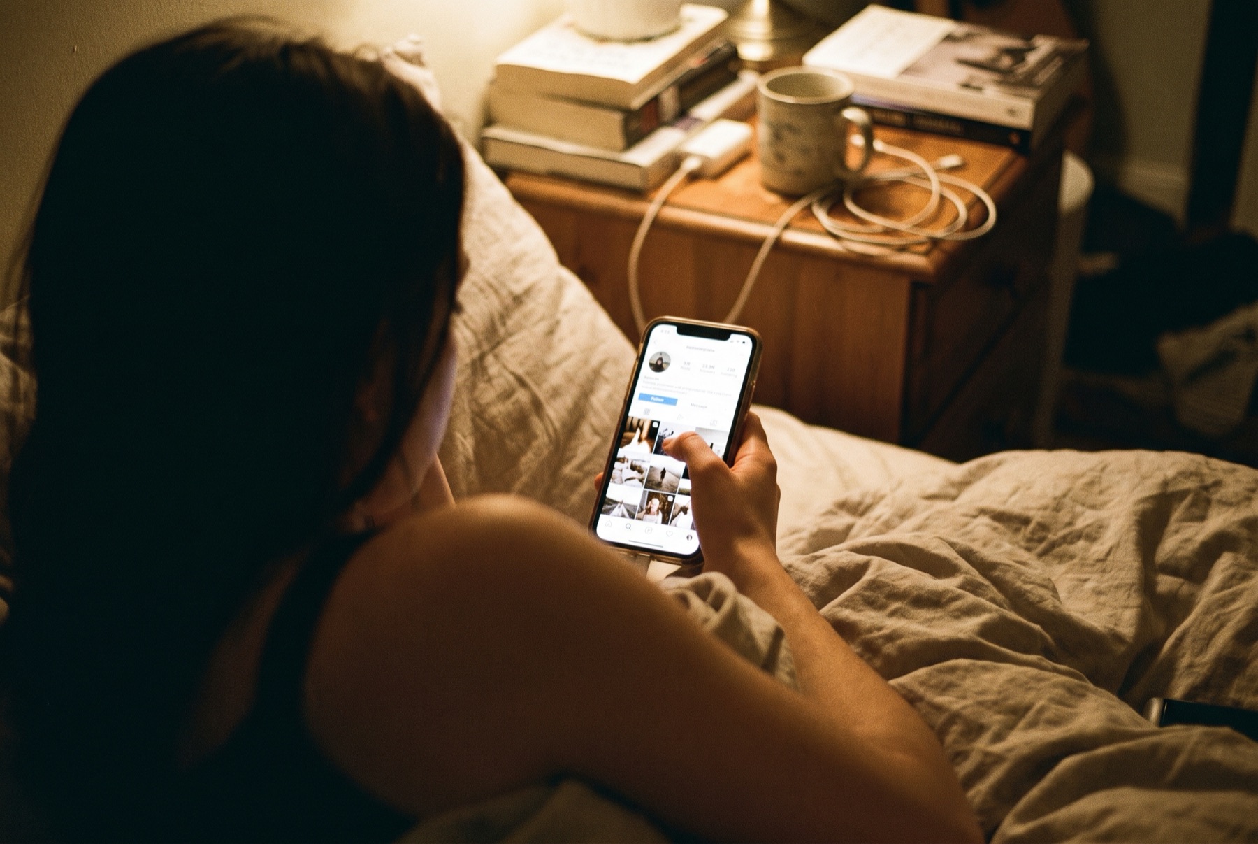 Over-the-shoulder view of a young woman scrolling on a phone in bed at night